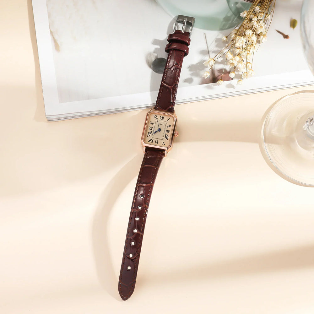 Rectangular gold wristwatch with brown leather strap on beige surface with magazine and dried flowers