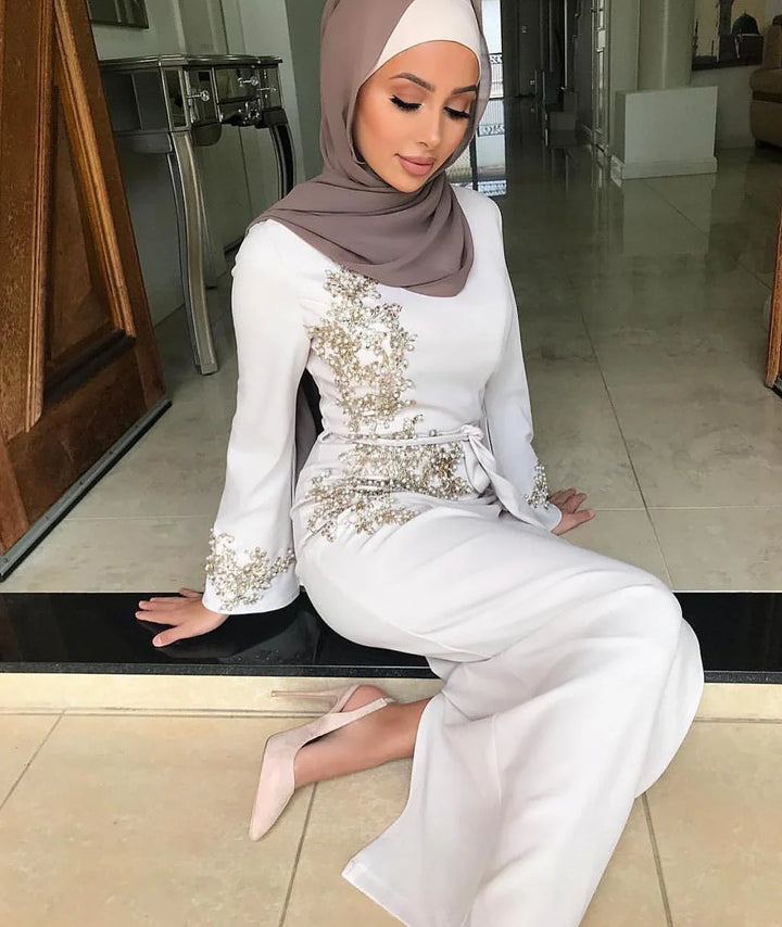 Woman wearing elegant white embellished dress and taupe hijab sitting indoors on tiled floor