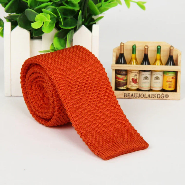 Rolled orange knitted tie on white surface with green plant and miniature wine bottle crate background