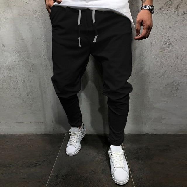 Man wearing black jogger pants with white drawstrings and white sneakers against gray wall