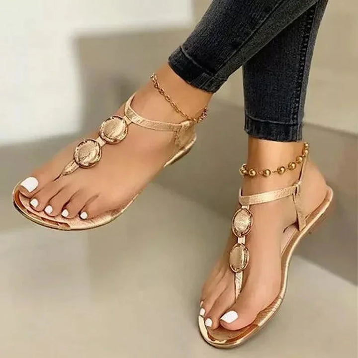Women's feet wearing gold flat sandals with round embellishments and ankle bracelets, paired with dark jeans