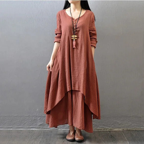 woman wearing rust-colored layered linen dress with long sleeves and pendant necklace, standing against gray wall