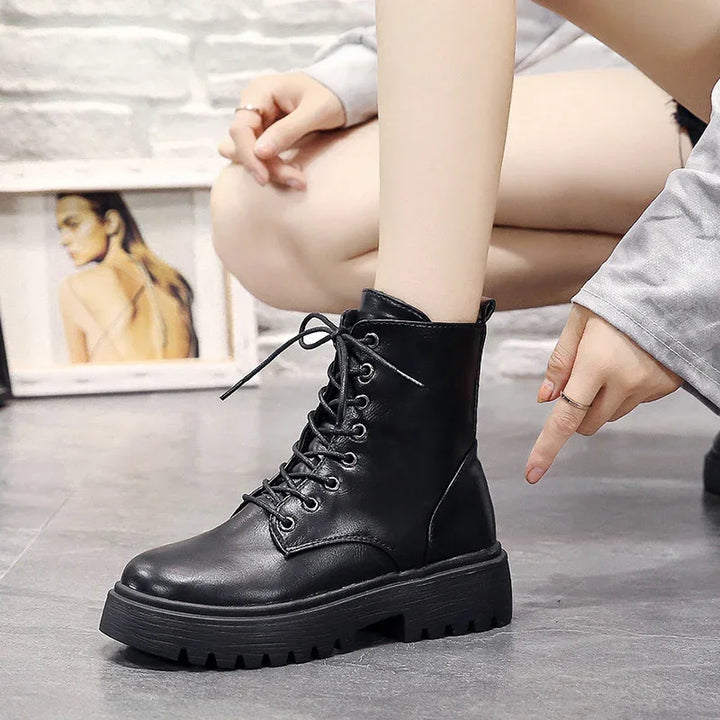 Black lace-up combat boot with thick rubber sole worn by person crouching on gray floor