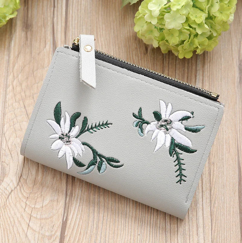 Light gray wallet with white floral embroidery and black zipper on wooden surface with green flowers