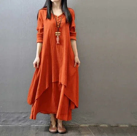 Woman wearing loose-fitting, layered burnt orange dress with long sleeves and brown shoes against gray wall