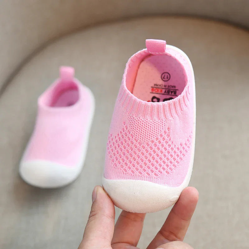 Hand holding pink baby sock shoes with white soles on beige fabric background