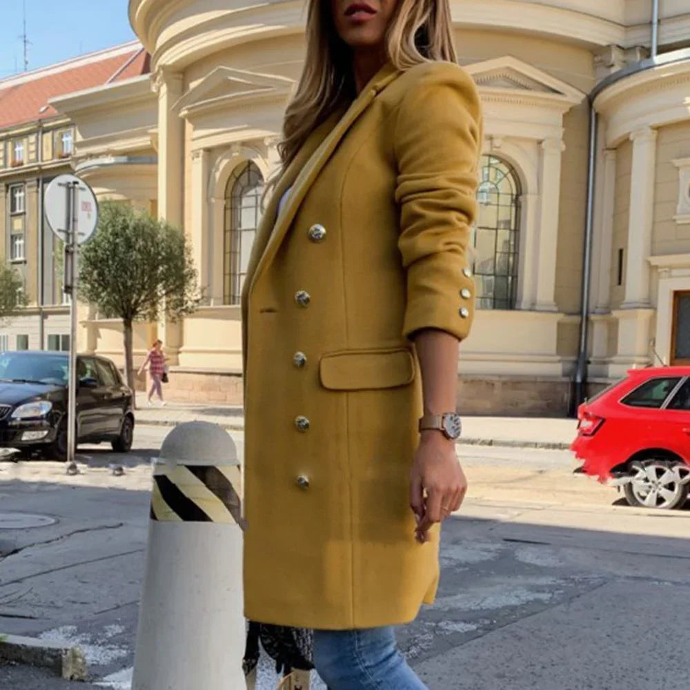 Woman wearing mustard yellow double-breasted coat and blue jeans standing on urban street