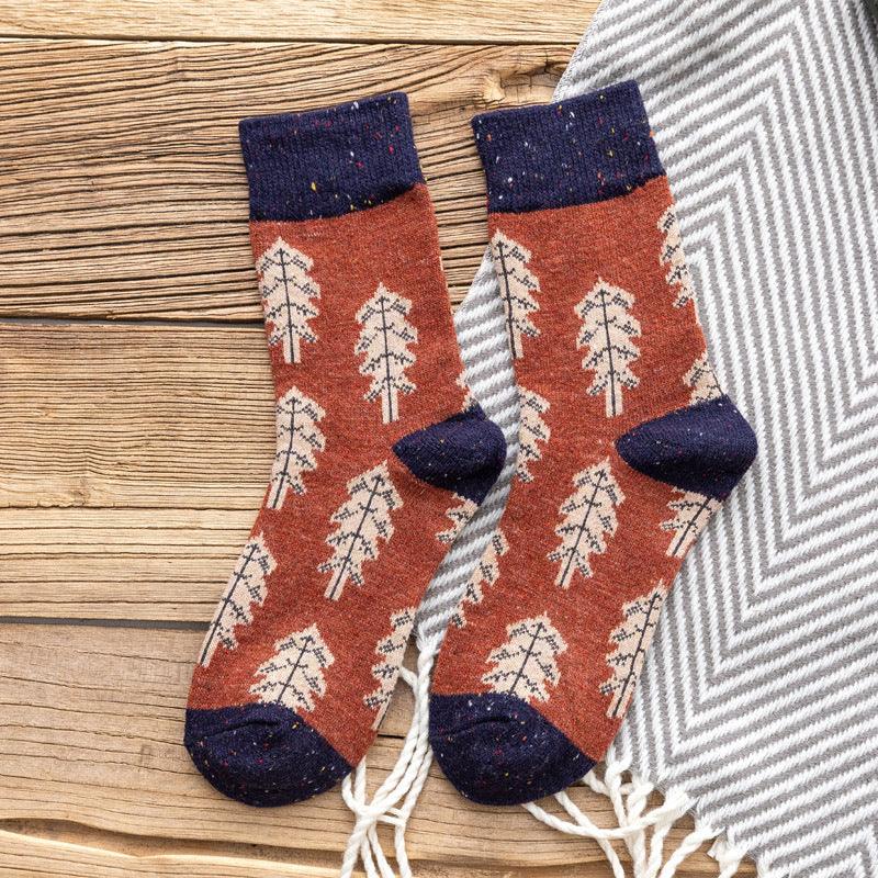 Pair of rust-colored socks with beige tree pattern and navy cuffs, heels, toes on wooden surface with gray chevron blanket