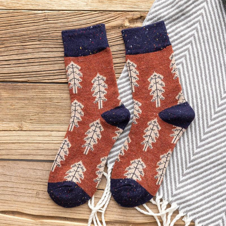 Pair of rust-colored socks with beige tree pattern and navy cuffs, heels, toes on wooden surface with gray chevron blanket