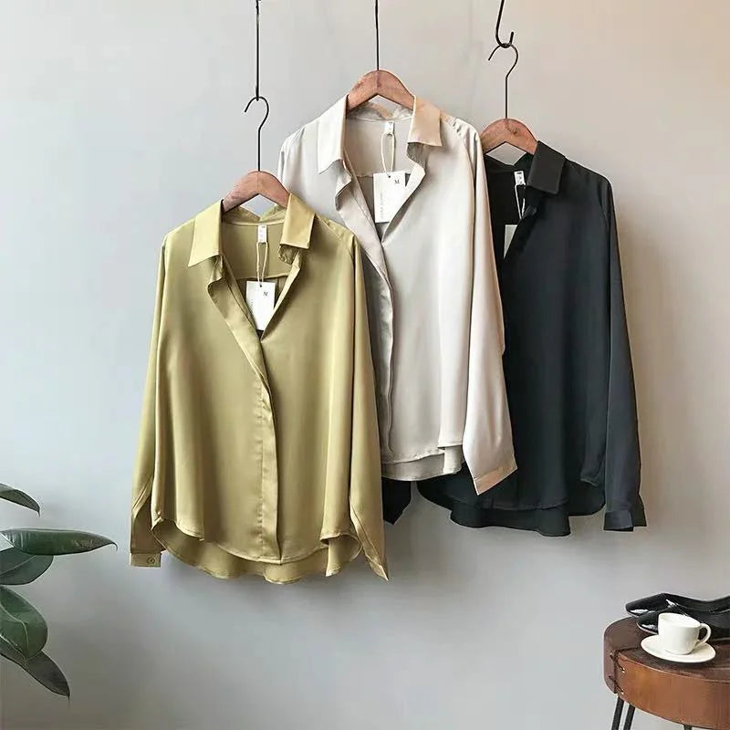 Three long-sleeve satin blouses in olive green, cream, and black hanging on wooden hangers against a light gray wall