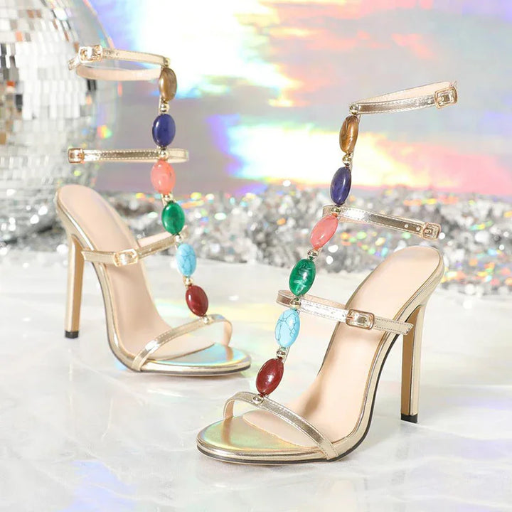 Gold high-heeled sandals with colorful oval gemstones and ankle straps on a reflective surface
