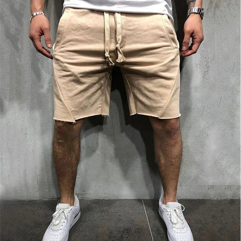 Man wearing beige drawstring shorts with raw hem and white sneakers against a gray wall