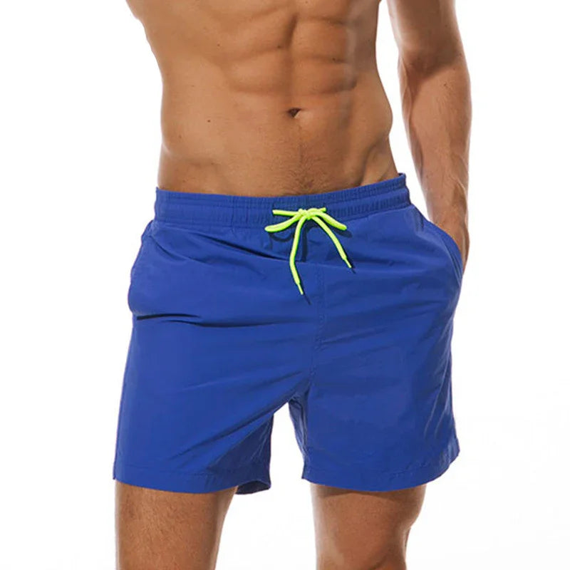 Man wearing blue swim shorts with neon green drawstring, hands in pockets, on white background