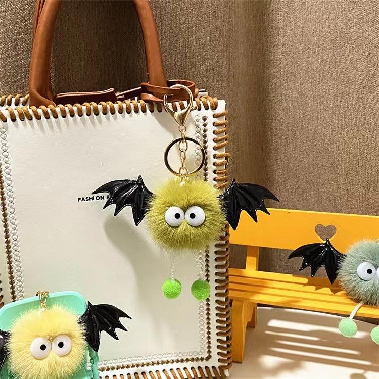 Furry green bat keychains with big eyes and black wings attached to white handbag and yellow bench