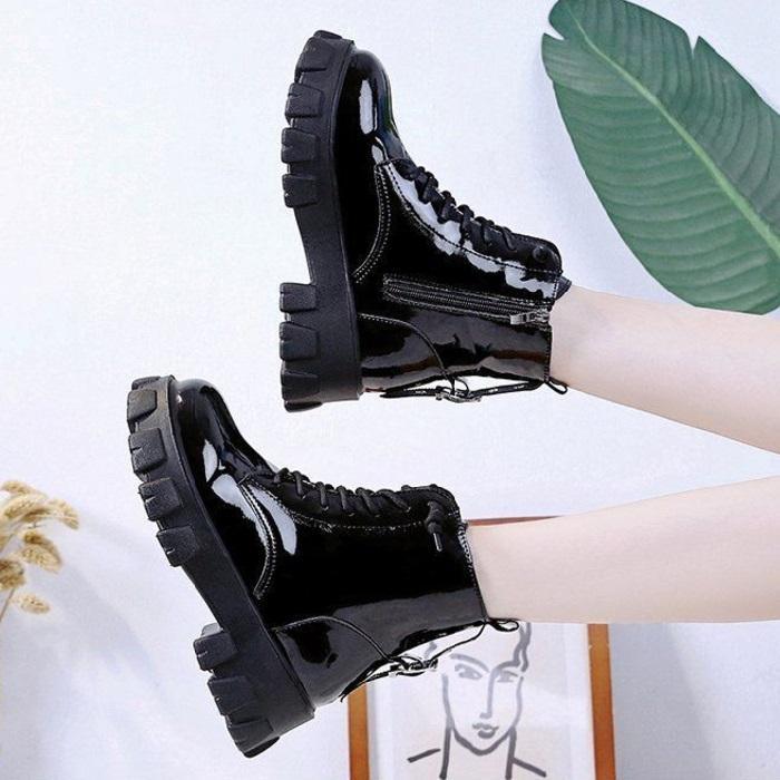 Shiny black patent leather combat boots with chunky soles worn on raised legs against a white wall