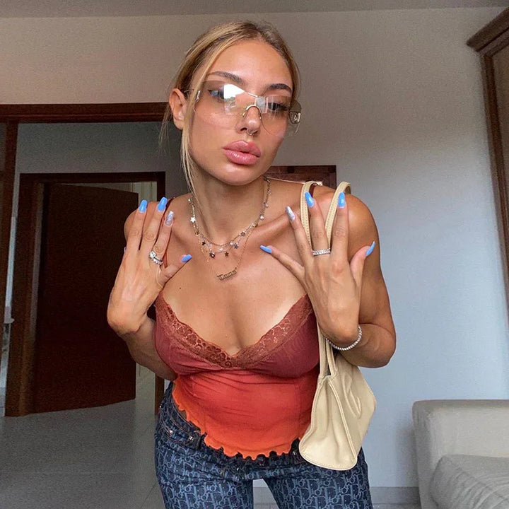 Young woman with blue nails wearing transparent glasses, orange lace cami top, and patterned jeans indoors