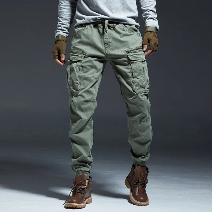 Man wearing green cargo pants with multiple pockets and brown leather boots on gray background