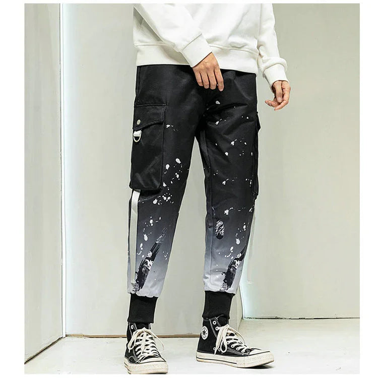 Men's black and gray gradient cargo pants with paint splatter and distressed details, paired with black high-top sneakers