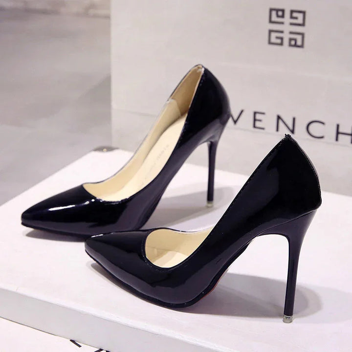 Pair of black patent leather high-heeled pumps on white surface with Givenchy bag in background