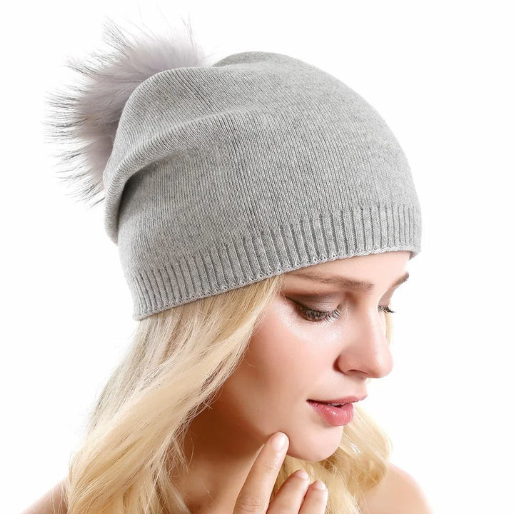 Blonde woman wearing light gray knit beanie hat with fluffy pom pom on white background