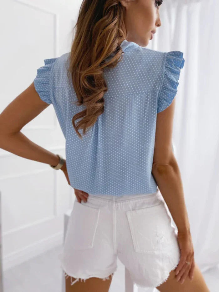 Woman wearing light blue polka dot ruffled sleeve blouse and white distressed denim shorts in bright room