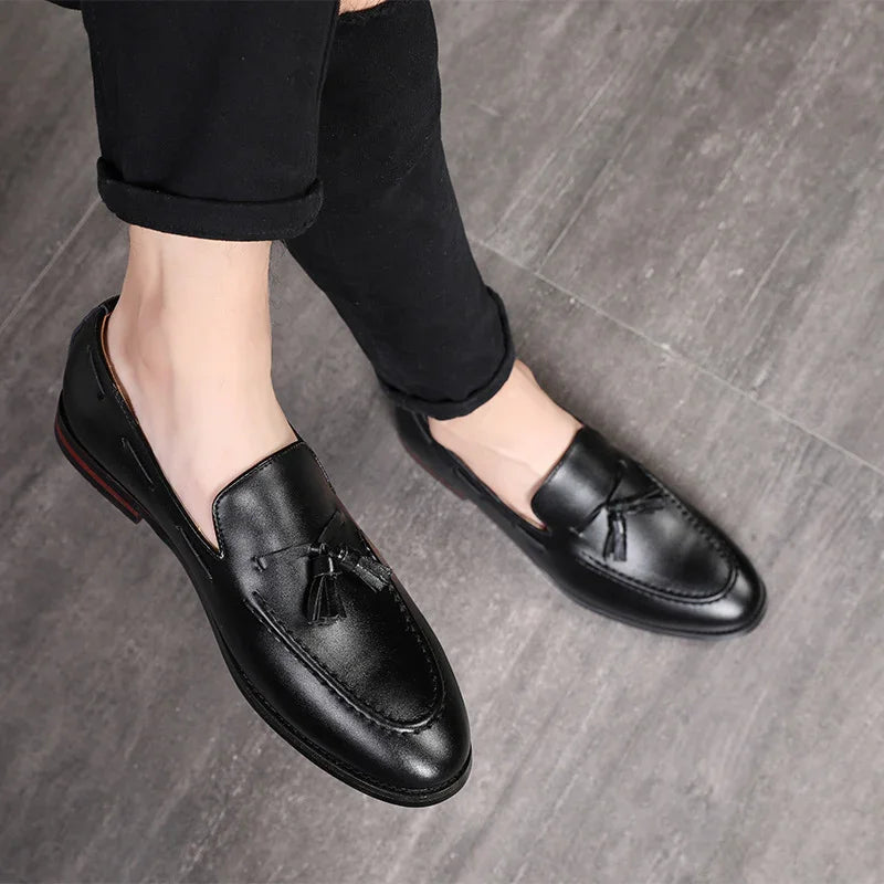 Black leather men's tassel loafers worn with rolled-up black pants on gray floor