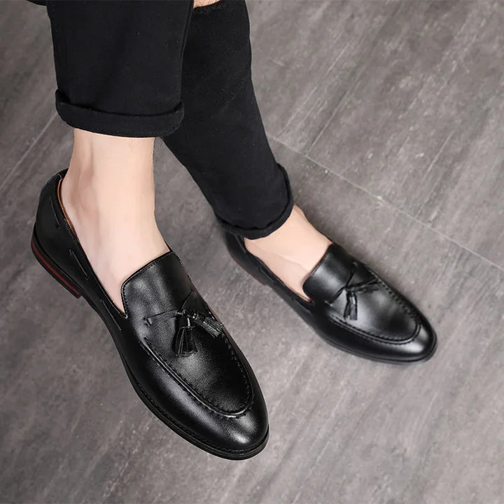 Black leather men's tassel loafers worn with rolled-up black pants on gray floor