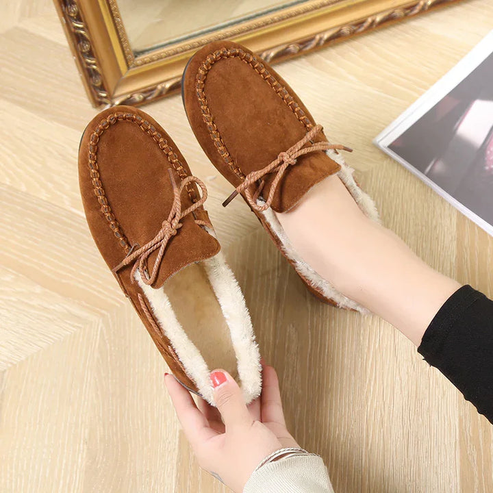 Brown suede moccasin slippers with fur lining and bow detail worn on feet on wood floor