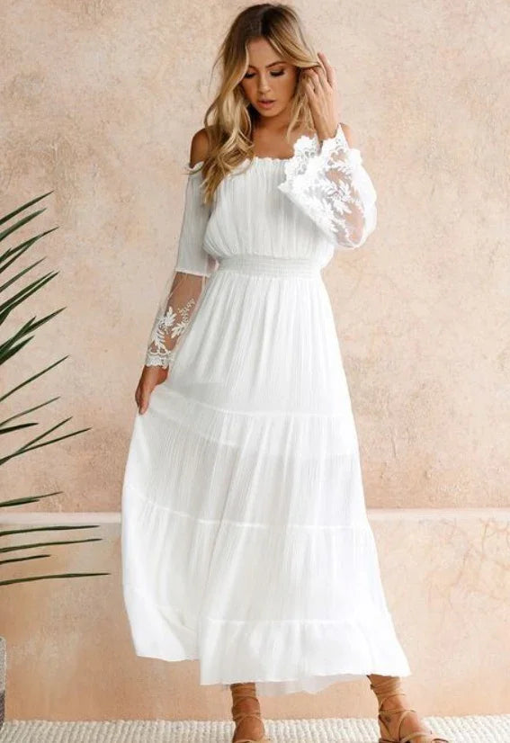 Woman wearing elegant white off-shoulder maxi dress with lace sleeves, standing against beige textured wall