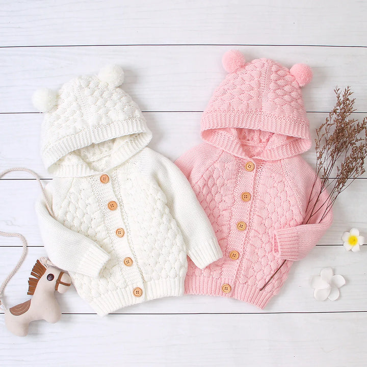 White and pink knitted baby hooded cardigans with pom pom ears and wooden buttons on white wooden background