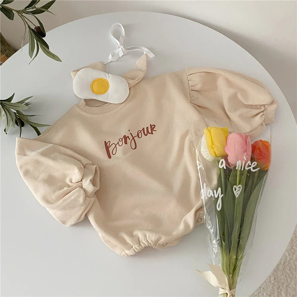 Beige baby romper with puff sleeves and 'Bonjour' text, paired with a fried egg eye mask and bouquet of tulips