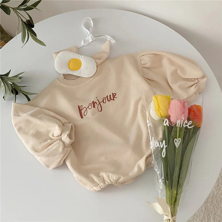 Beige baby romper with puff sleeves and 'Bonjour' text, paired with a fried egg eye mask and bouquet of tulips