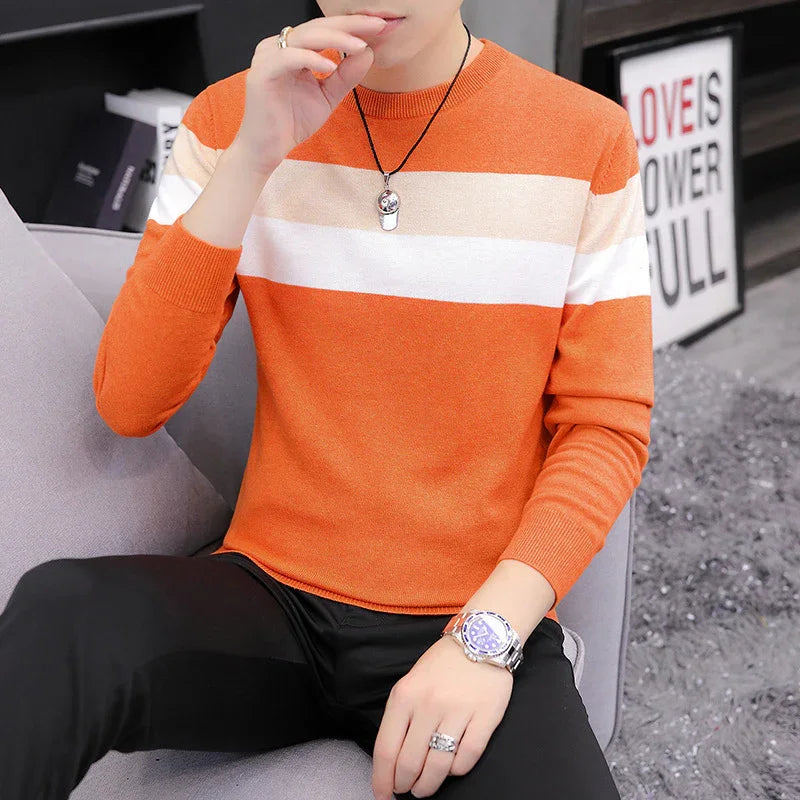 Man wearing orange and white striped sweater, black pants, silver watch, and a pendant necklace sitting on a gray chair