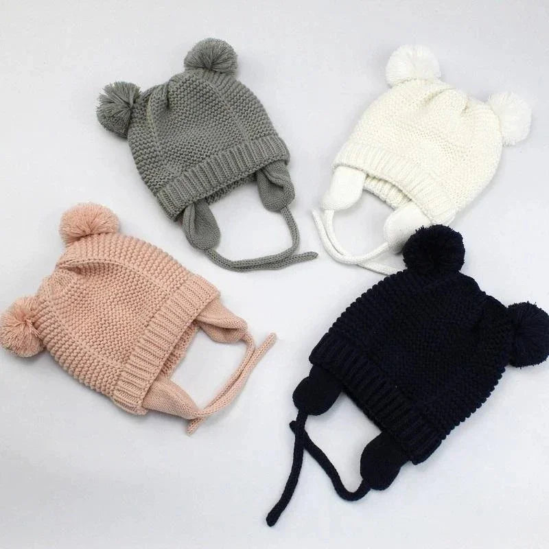 Four knitted baby hats with ear flaps and pom-poms in gray, ivory, pink, and navy colors