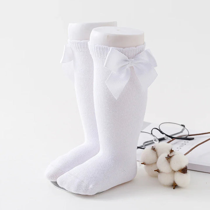 Pair of white knee-high baby socks with decorative bows on mannequin legs
