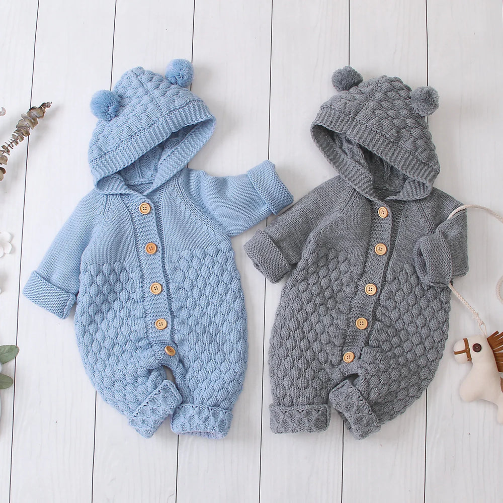 Two knitted baby rompers with hoods and pom-poms in blue and gray on white wooden background