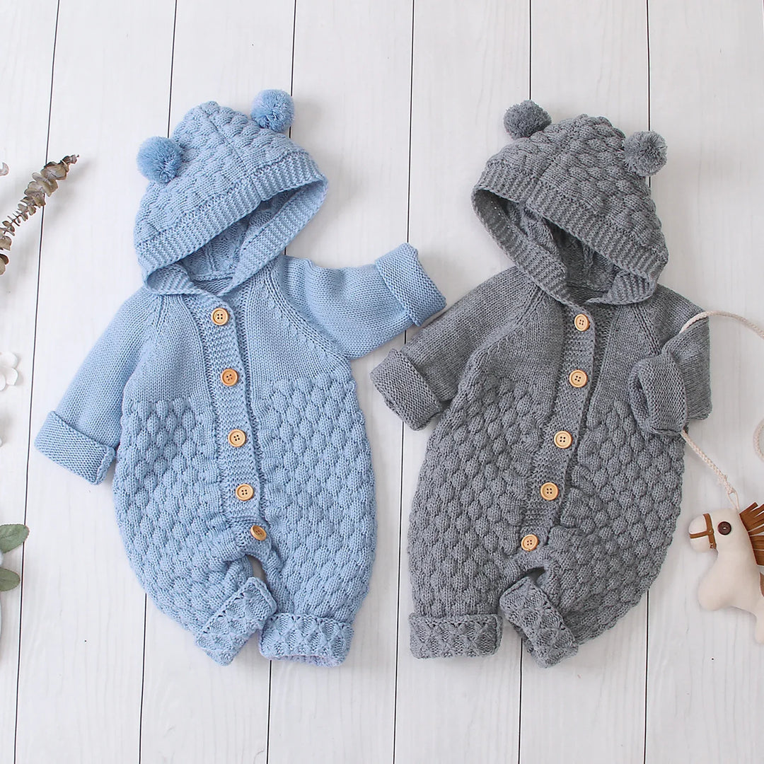 Two knitted baby rompers with hoods and pom-poms in blue and gray on white wooden background