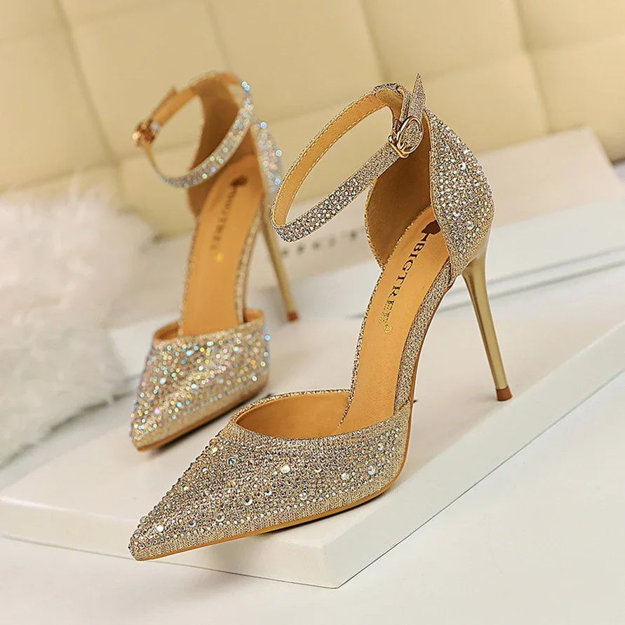 Elegant gold high-heeled shoes with sparkling rhinestones and ankle straps on white surface
