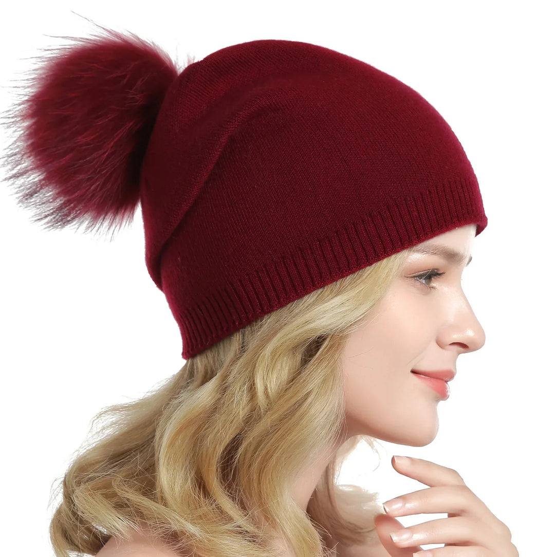 Profile of woman wearing burgundy knit beanie hat with large fluffy pom-pom, blonde hair, white background