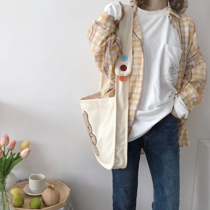 Person wearing yellow plaid shirt over white graphic tee with denim jeans and canvas tote bag with colorful buttons