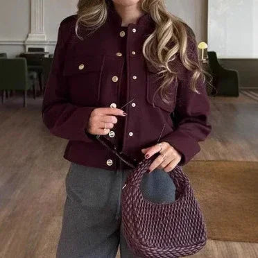 Woman wearing a stylish purple buttoned jacket and grey pants holding a woven purple handbag indoors