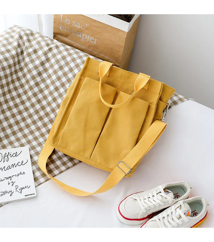 Yellow canvas tote bag with front pockets and adjustable strap on checkered fabric and white surface with sneakers