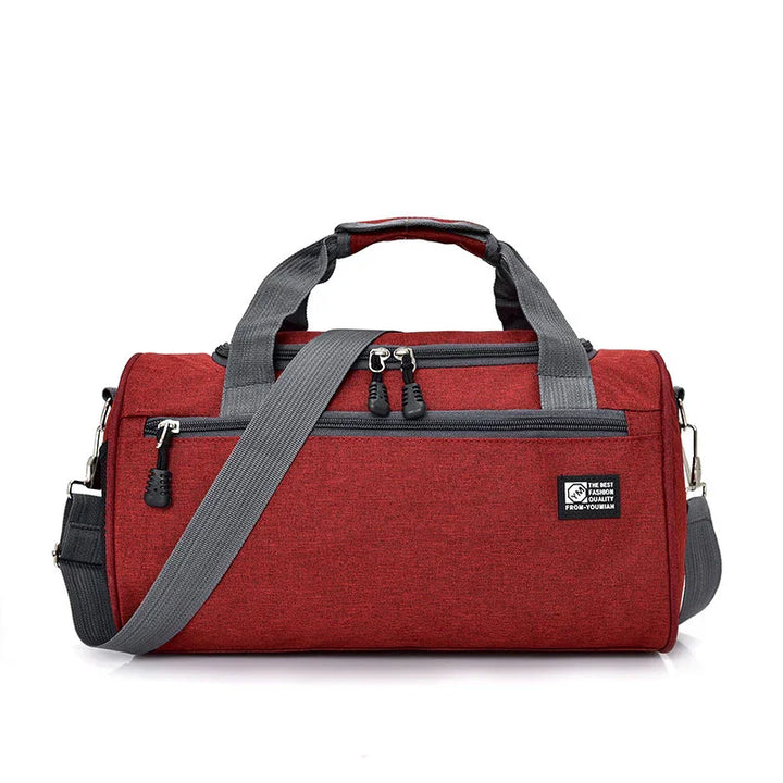 Red duffel bag with gray handles and shoulder strap, multiple zippered pockets, and black logo patch