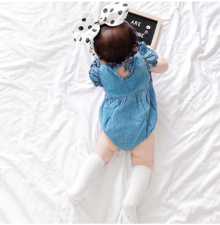 Baby wearing denim romper, white knee-high socks, and polka dot bow headband lying on white bed with message board