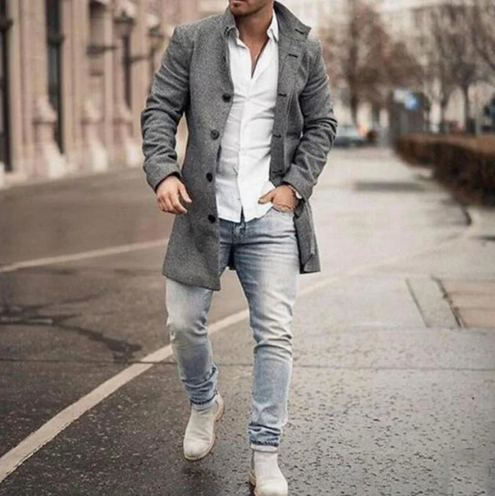 Man walking on city street wearing grey buttoned coat, white shirt, light blue jeans, and beige shoes