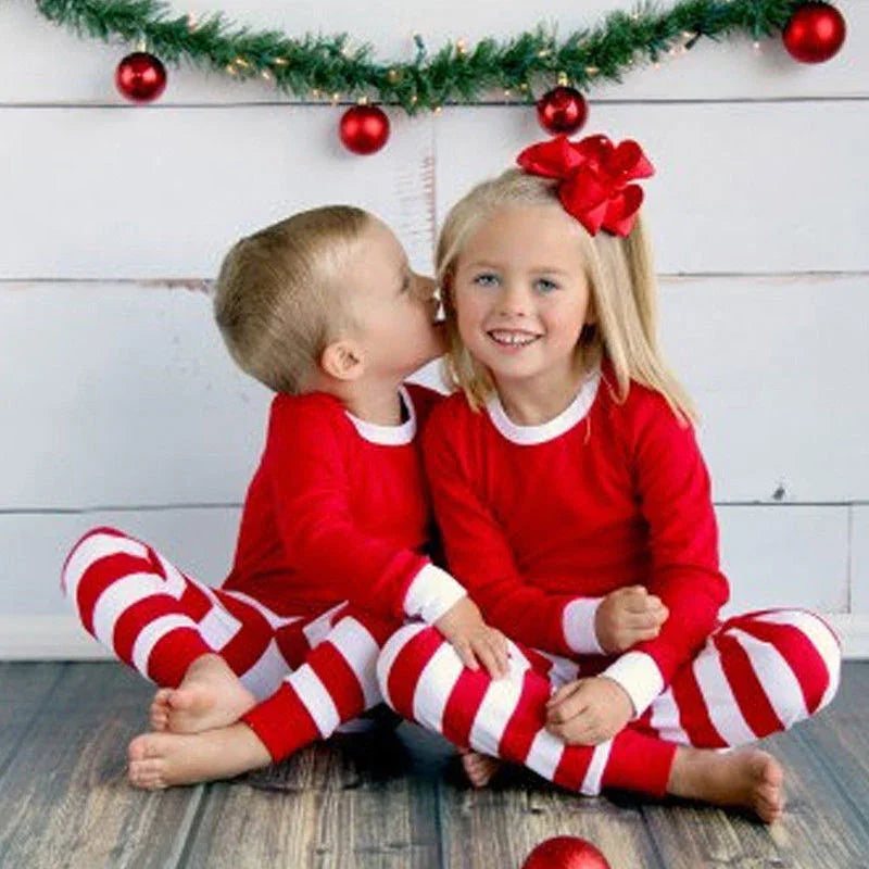 Two children in matching red and white striped Christmas pajamas sitting on wooden floor with holiday garland and red ornaments