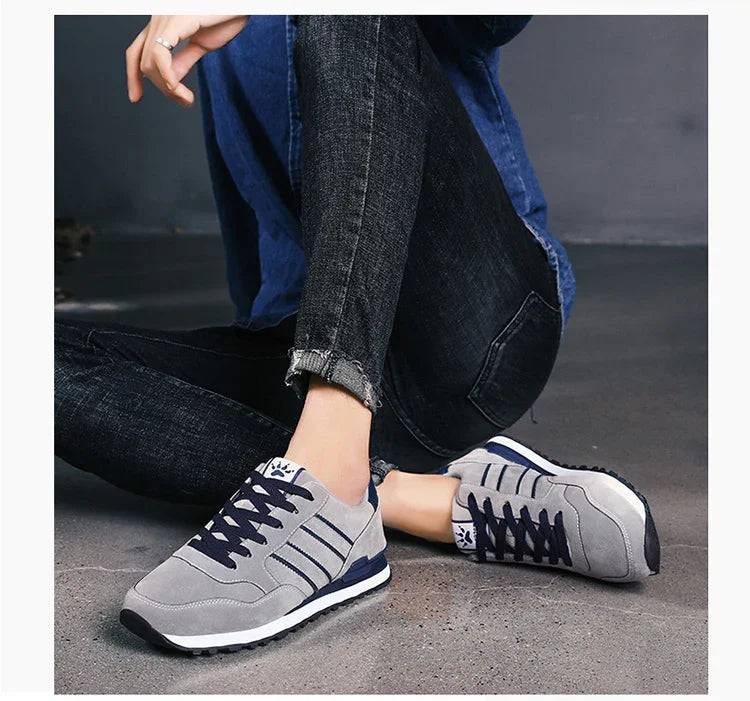 Person wearing gray and navy casual sneakers with dark jeans sitting on gray floor against dark wall
