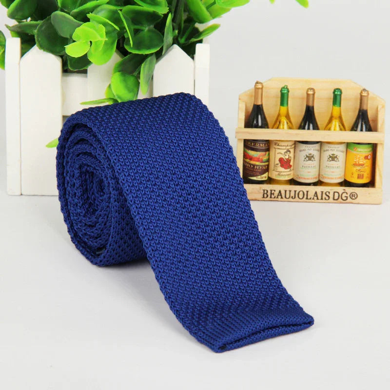 Rolled blue knitted necktie on a white surface with green plant and miniature wine bottles in wooden crate