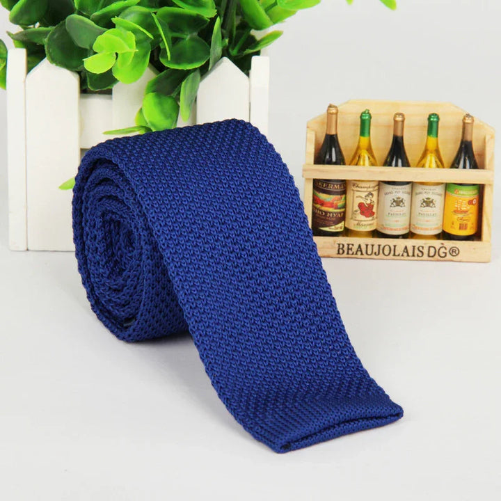 Rolled blue knitted necktie on a white surface with green plant and miniature wine bottles in wooden crate
