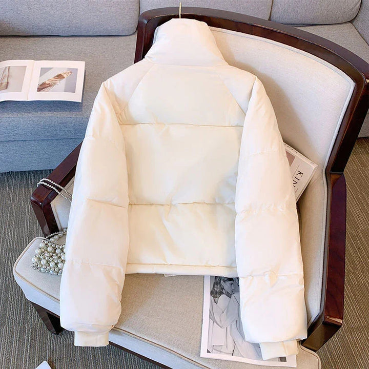 Cream white puffer jacket with high collar laid on beige armchair with magazine and pearl bag